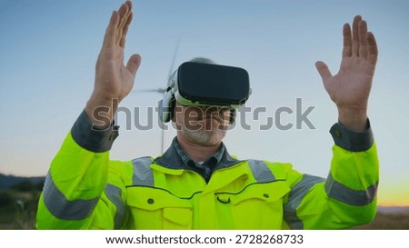 Engineer in high visibility jacket using VR headset outdoors. Man lifting both hands while interacting with virtual interface. Professional applying virtual reality at wind energy project site.