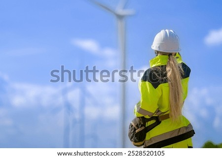 An engineer in high visibility work gear and a hard hat points toward a wind turbine in the background. The scene highlights renewable energy and the professional's involvement in wind farmoperations.
