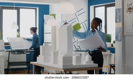 Engineer holding blueprints plan and analyzing building model for architecture work. Woman architect working with tools to design construction layout and structure for urban project.