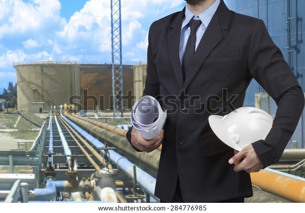 Engineer\
holding hard hat and blueprint for working at pipe line connection\
to oil tanks in petrochemical oil\
refinery