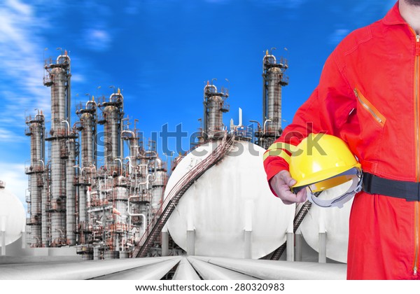 Engineer\
holding hard hat for working at pipe line connection to oil tanks\
in petrochemical oil refinery against blue sky\
