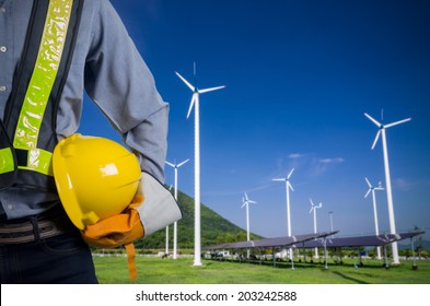 Engineer holding a yellow helmet. Against a backdrop of renewable energy farms.
