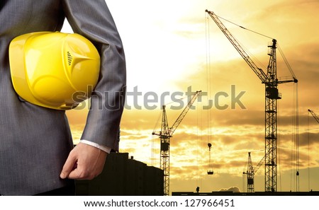 engineer holding yellow helmet for workers security on background of  new highrise apartment buildings and construction cranes on background of evening sunset cloudy sky Silhouette Crane lifts load