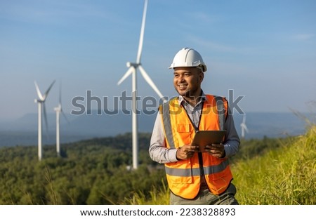Engineer India man working with tablet at windmill farm Generating electricity clean energy. Wind turbine farm generator by alternative green energy. Asian engineer checking control electric power
