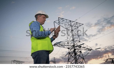 Engineer inputs data on tablet and nods head by power transmission lines support