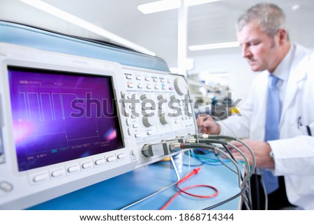 An Engineer in lab coat working at an electrical test bench next to an oscilloscope in selective focus