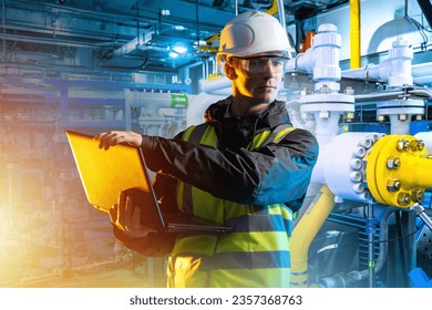 Engineer with laptop. Man with computer. Engineer in basement of industrial building. Industrialist man in protective helmet. Factory employee. Gas pipes behind guy. Chemical factory engineer