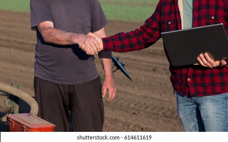 Engineer with laptop shaking hands with farmer beside sowing equipment on tractor in the field in spring
