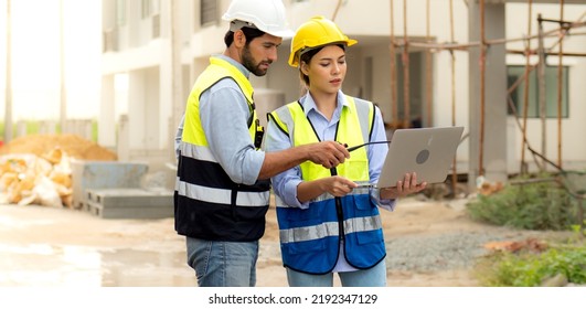 Engineer Man And Female Architect Wear Safety Helmets Discuss Housing Development Project At Construction Site Using Laptop Computer. Contractor Manager Examining Building Estate Infrastructure.