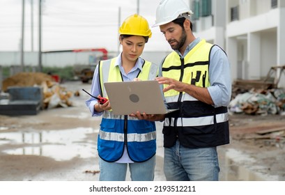 Engineer Man And Female Architect Wear Safety Helmets Discuss Housing Development Project At Construction Site Using Laptop Computer. Contractor Manager Examining Building Estate Infrastructure.