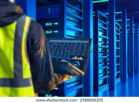 IT engineer. Man with laptop in server room. Monitoring load on IT infrastructure. System administrator in yellow vest. IT engineer with back to camera. Hosting server maintenance.