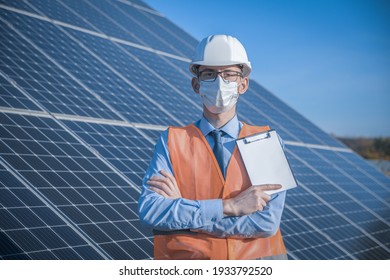 Engineer, man in uniform and mask, helmet glasses and work jacket on a background of solar panels at solar station. Technician check the maintenance