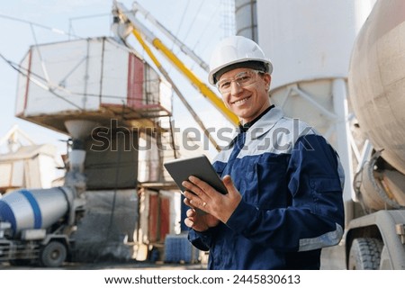 Engineer man in uniform with tablet computer controls loading of cement to mixer truck on concrete factory. Concept automated logistics on industry with online internet.
