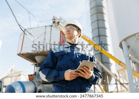 Engineer man in uniform with tablet computer controls loading of cement to mixer truck on factory. Concept banner automated logistics online internet.