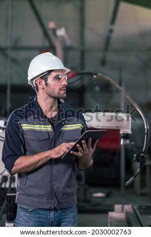 Engineer man use tablet to control welding robotic automatic arm machine in the smart factory. Looking at camera. Automation manufacturing for industrial 4.0. Technology, software and safety control