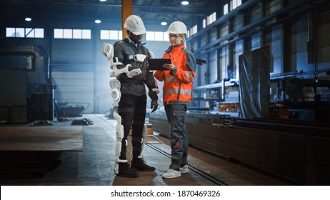 Engineer And Manual Labor Worker In A Bionic Exoskeleton Prototype Check Development Test Results On A Tablet. Heavy Industry Factory Engineers Discuss Warehouse Work And Futuristic Technology.