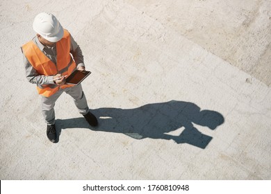 An engineer in an orange vest and a white construction control helmet conducts an inspection with a tablet in his hands, top view
