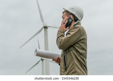 Engineer with a plan in hand talking on a mobile phone near wind generators in cloudy weather