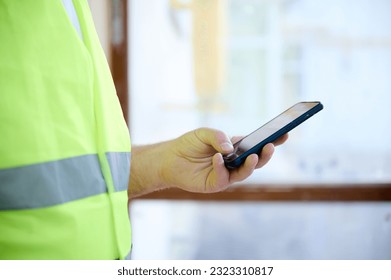 Engineer in protective vest using his smart phone at the construction site.Crop portrait of mature construction worker in uniform browsing social media on mobile phone during break. Crop view. - Powered by Shutterstock