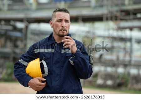 Engineer or refinery worker holding hardhat smoking at construction site outdoor with background of petroleum industry. Pipeline transport production line