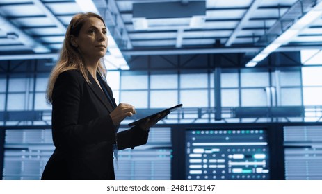 Engineer in server hub checking recovery plan on tablet, monitoring rackmounts energy consumption. Woman in data center making sure rigs are functioning optimally, doing routine disk checks, camera A