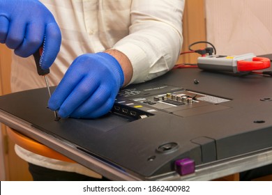 Engineer At The Service Center Removes Back Panel From TV Monitor LED For Repair. Gloved Hands Hold Screwdriver. Close-up, Selective Focus, Blur.