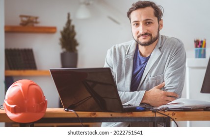 The engineer smiles and looks in front of him working with drawings and computers at the table on which the helmet lies. Color graded