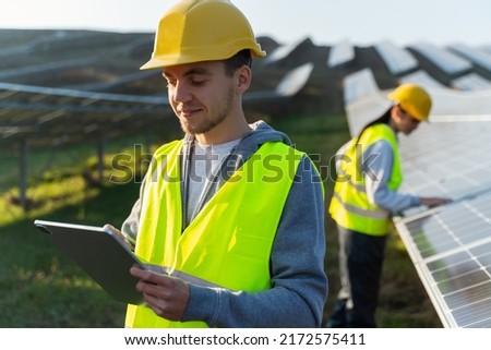 Engineer of solar power plant blogging on company page on social network. Young man in yellow vest holding tablet in hands, posting new content on background of solar panels 