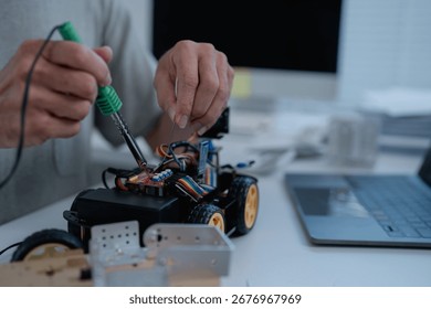 Engineer is soldering electronic components of a robot car prototype in a laboratory using a soldering iron and tin wire, with a laptop and documents in the background - Powered by Shutterstock - Get 15% off with code: PIKWIZARD15