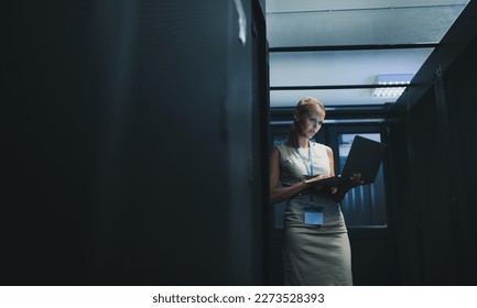IT engineer standing amid working server racks doing routine maintenance check and diagnostics using laptop computer (color toned image)
