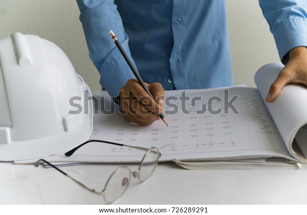 engineer standing behind the table to write on the
paper near by the white safety cap on and there are glasses on the
table blurry