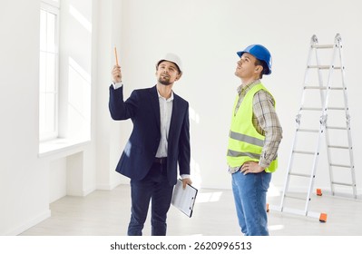 Engineer standing with builder in empty apartment room with ladder in background and pointing at something. Architect or designer consulting foreman in making renovation and design of living space.