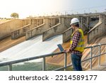 Aืn Engineer standing by a dam. He is wearing a white hard hat and yellow transparent vest. He is doing this daily inspection work.