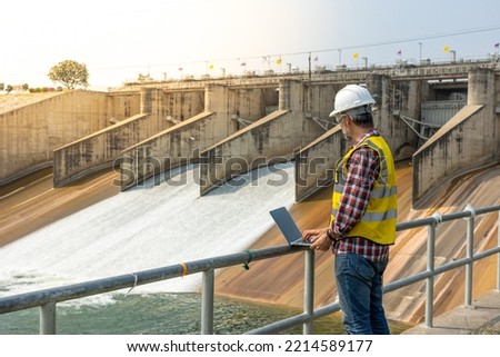 Aืn Engineer standing by a dam. He is wearing a white hard hat and yellow transparent vest. He is doing this daily inspection work.