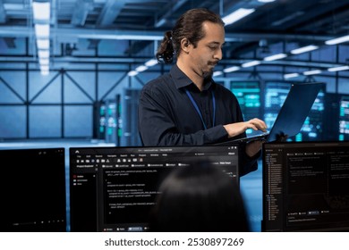 Engineer standing in data center, typing on laptop, doing yearly checkup to avoid equipment downtime. IT employee in server hub monitoring server infrastructure components using tablet