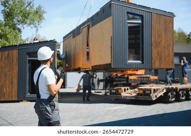 Engineer supervises the loading of a prefabricated modular house onto a truck, ensuring efficient and safe transportation to its destination