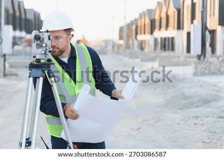 Engineer or surveyor using a total station instrument while holding blueprints at a developing housing construction site