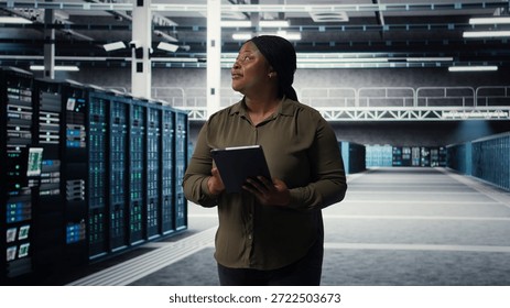 Engineer with tablet walks through data center server rows, implementing advanced power management solutions. IT professional using device to oversee server hub, reducing energy consumption, camera A