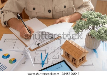 Engineer is taking notes on energy consumption data while designing a sustainable house powered by renewable energy, promoting an ecological future