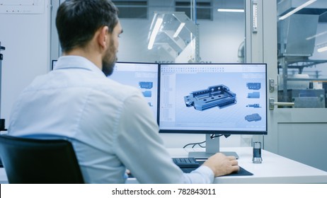 Engineer/ Technician Working on a Personal Computer with Two Displays, He's Designing New Component in CAD Program. Out of the Office Window Components Manufacturing Factory is Seen. - Shutterstock ID 782843011
