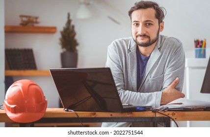 The engineer is thinking and smiling while working with drawings and computers at the table on which the helmet lies. Color graded
