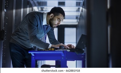 IT Engineer with Tool Cart Working on a Laptop Computer, he Deploys new software. He Stands at a Corridor of a Large Data Center Full of Rack Servers. - Shutterstock ID 662881918