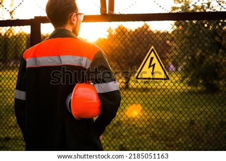 an engineer in uniform with a safety helmet stands at the entrance to a power plant with a danger sign on the fence