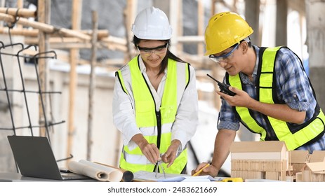 An engineer uses a compass on blueprints while meeting with a foreman at a construction site, Discuss project details, and are surrounded by tools, a laptop, and a scale model. - Powered by Shutterstock