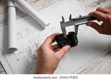 Engineer using caliper measuring prototype part on blueprint in design office.