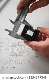 Engineer using caliper measuring prototype part on blueprint in design office.