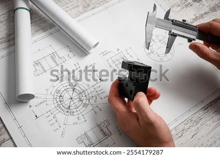 Engineer using caliper measuring prototype part on blueprint in design office.