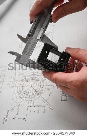 Engineer using caliper measuring prototype part on blueprint in design office.