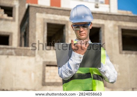 Engineer using laptop on construction site wearing safety helmet and vest. Mobile technology and modern construction business solutions.