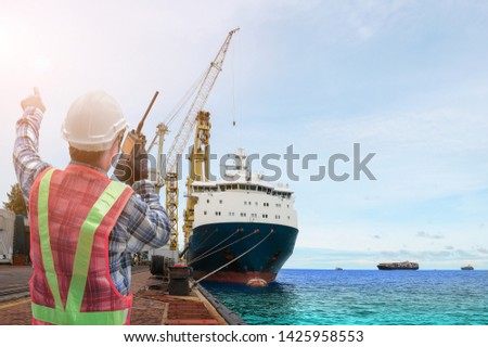 Engineer with walkie talkie in hand holding or portable radio transceiver for communication in shipping port export nautical vessel transportation and industry logistic and container ship sailing.